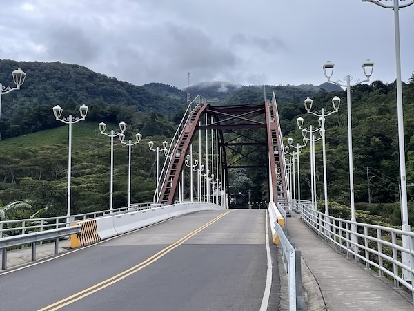 A metal bridge with forested hills and clouds behind it.