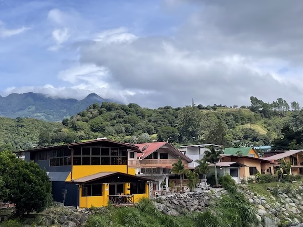 A row of houses next to a river with mountains, forest and clouds in the background.