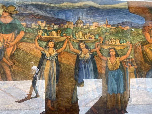 A mural showing women carrying baskets on their head, a walking man is reflected in the glass.