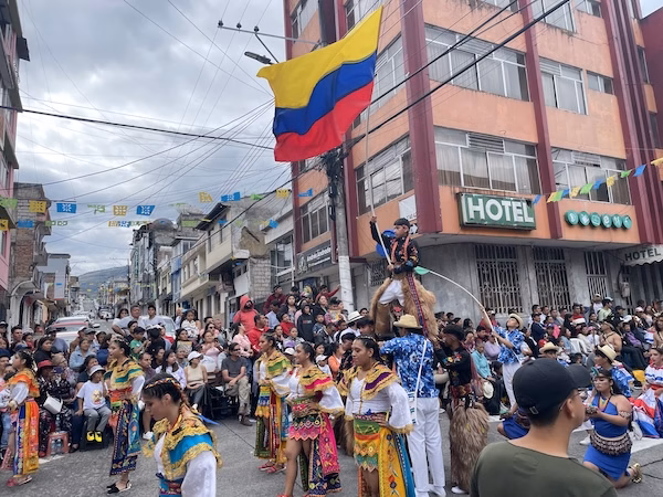 A boy in traditional chaps waves a yellow, blue and red flag while surrounded by dancers in traditional dresses during a parade.